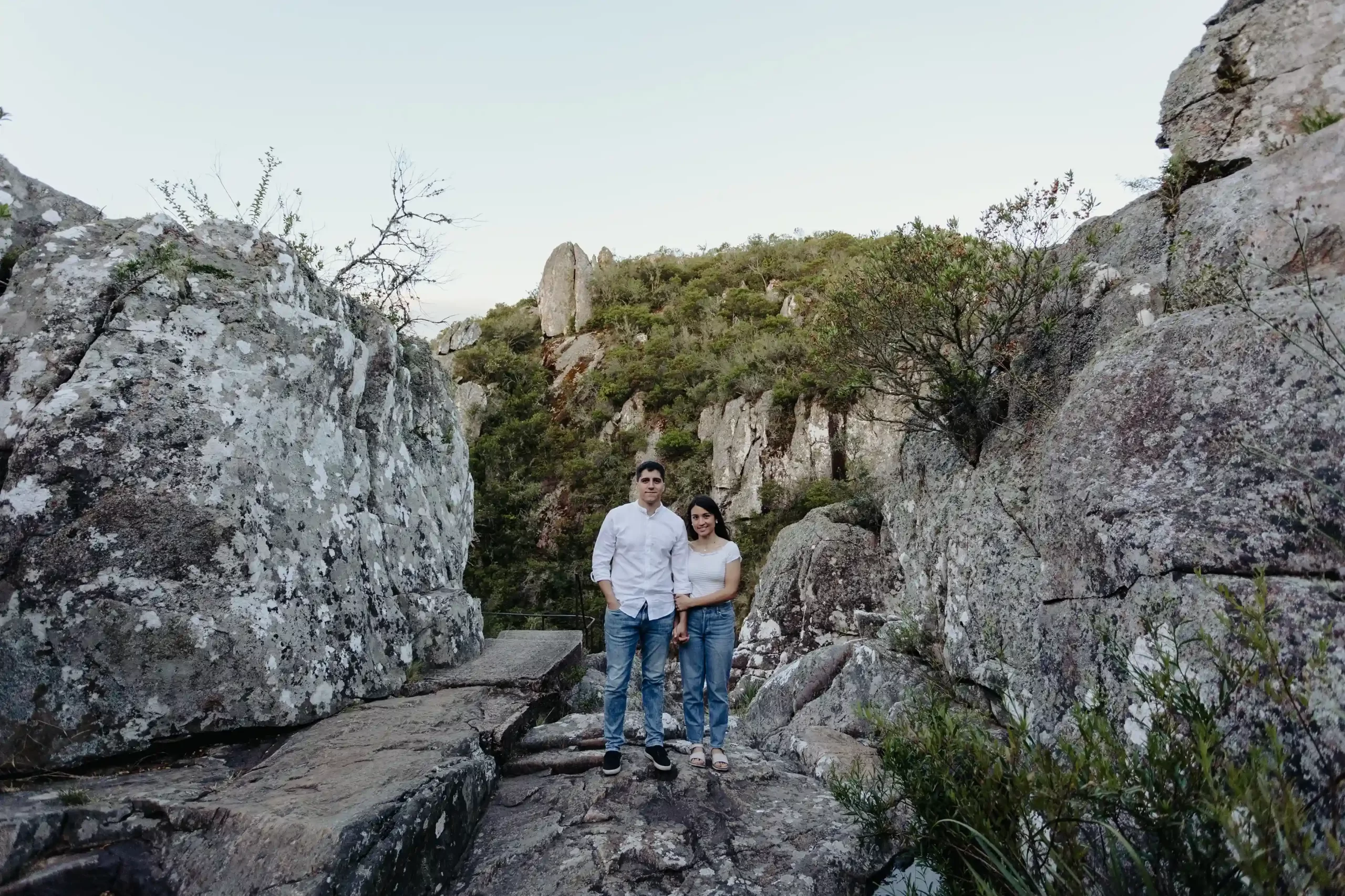 Pareja de pie juntos en un sendero rocoso rodeado de formaciones rocosas y vegetación en un paisaje montañoso al atardecer, capturados por fotógrafos de boda en Uruguay