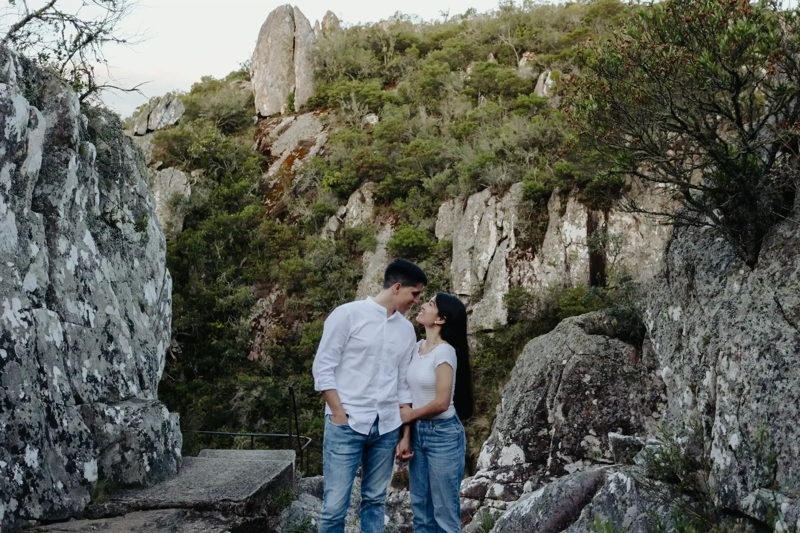 Pareja de pie juntos en un sendero rocoso rodeado de formaciones rocosas y vegetación en un paisaje montañoso al atardecer, capturados por fotógrafos de boda en Uruguay