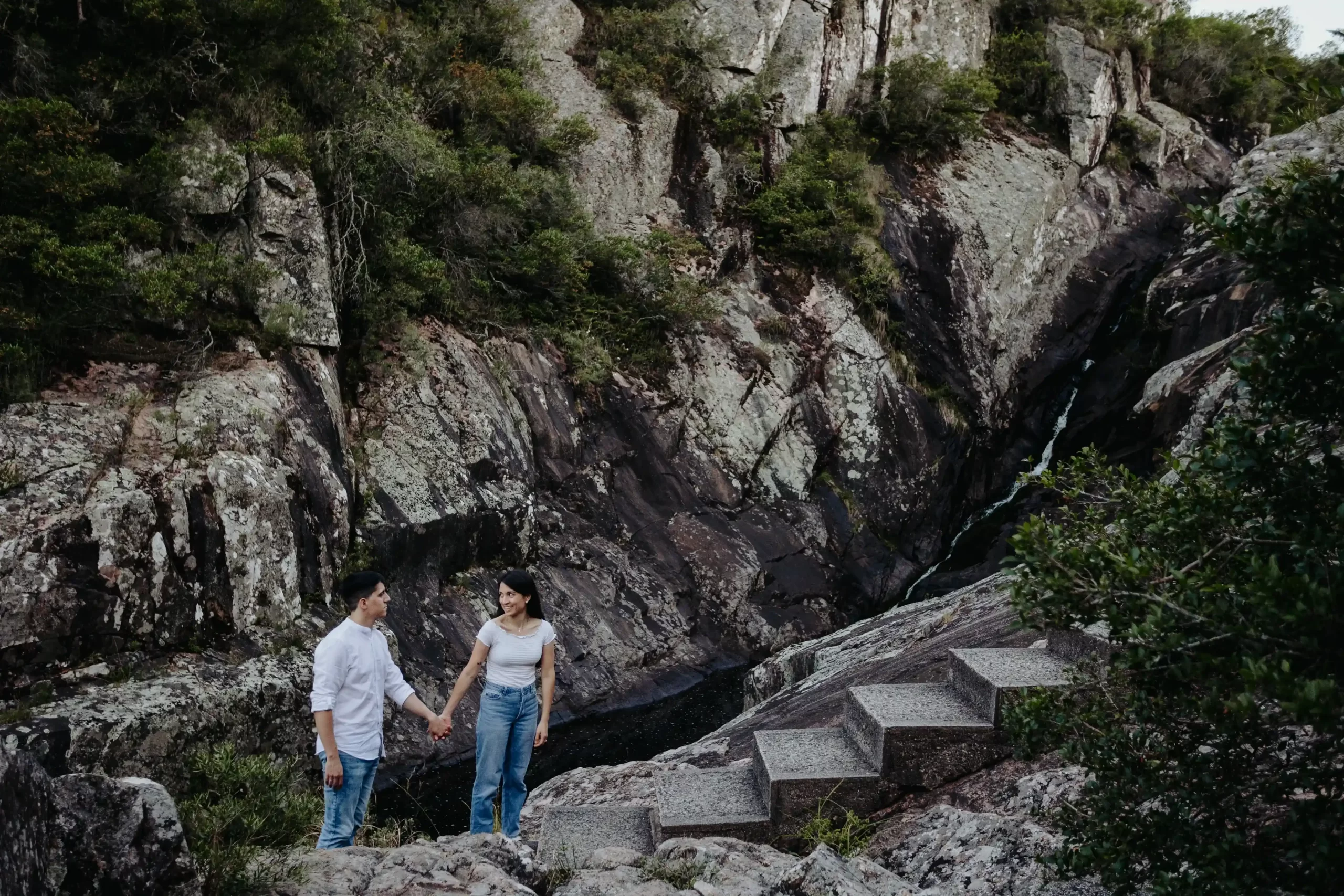 Pareja de pie juntos en un sendero rocoso rodeado de formaciones rocosas y vegetación en un paisaje montañoso al atardecer, capturados por fotógrafos de boda en Uruguay