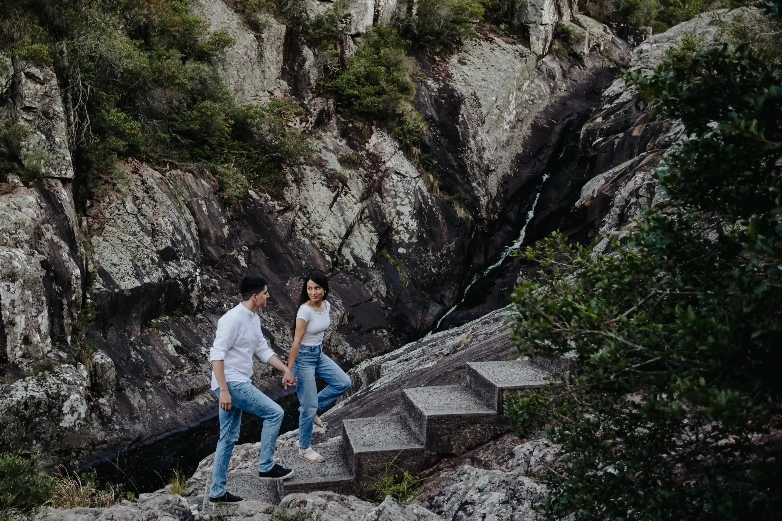 Pareja de pie juntos en un sendero rocoso rodeado de formaciones rocosas y vegetación en un paisaje montañoso al atardecer, capturados por fotógrafos de boda en Uruguay